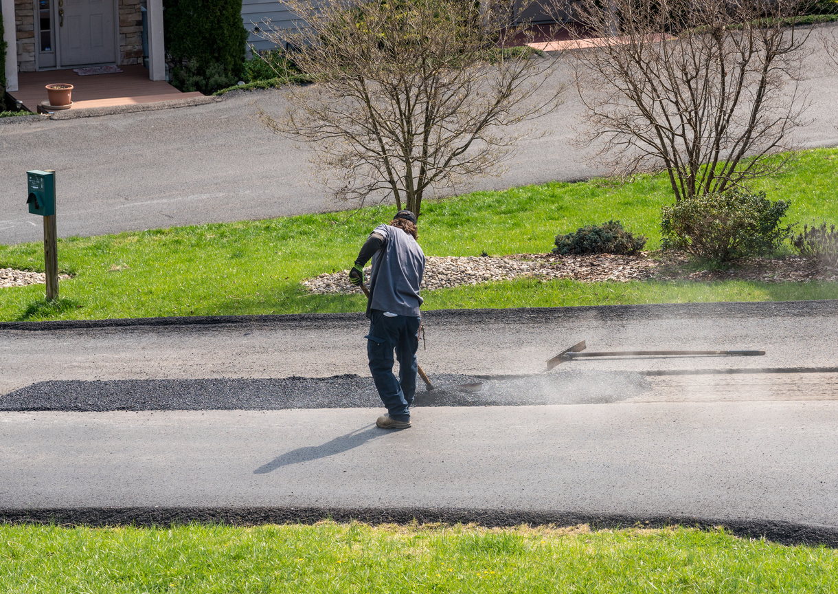172254365681472090 (44) Fall Asphalt Maintenance Worker using a machine to resurface a road in a suburban area with greenery and houses in the background.