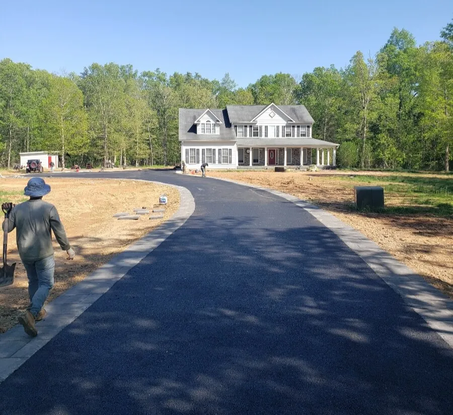 A person walks on a newly paved driveway leading to a two-story house with white siding, surrounded by trees.