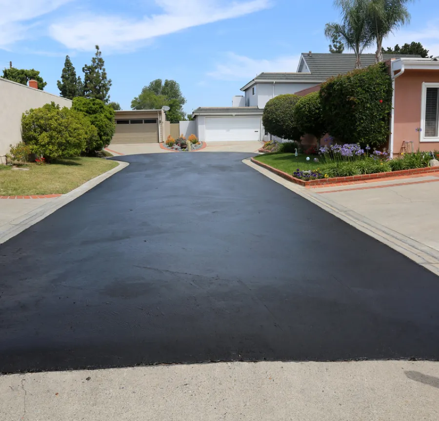 Freshly paved residential driveway with surrounding greenery and houses in the background under a clear sky.