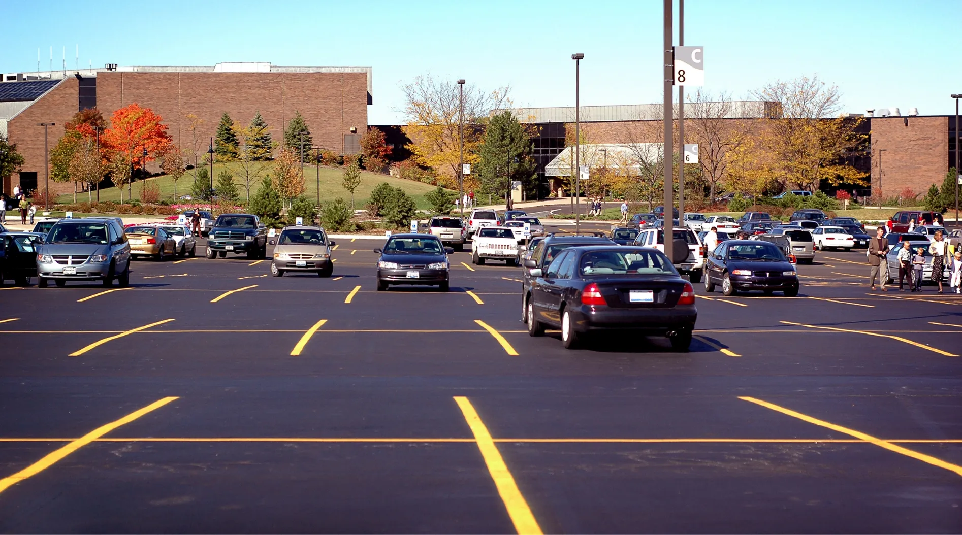 parking-lot-1 Cars parked in a spacious parking lot with a few vehicles moving, surrounded by buildings and autumn trees.