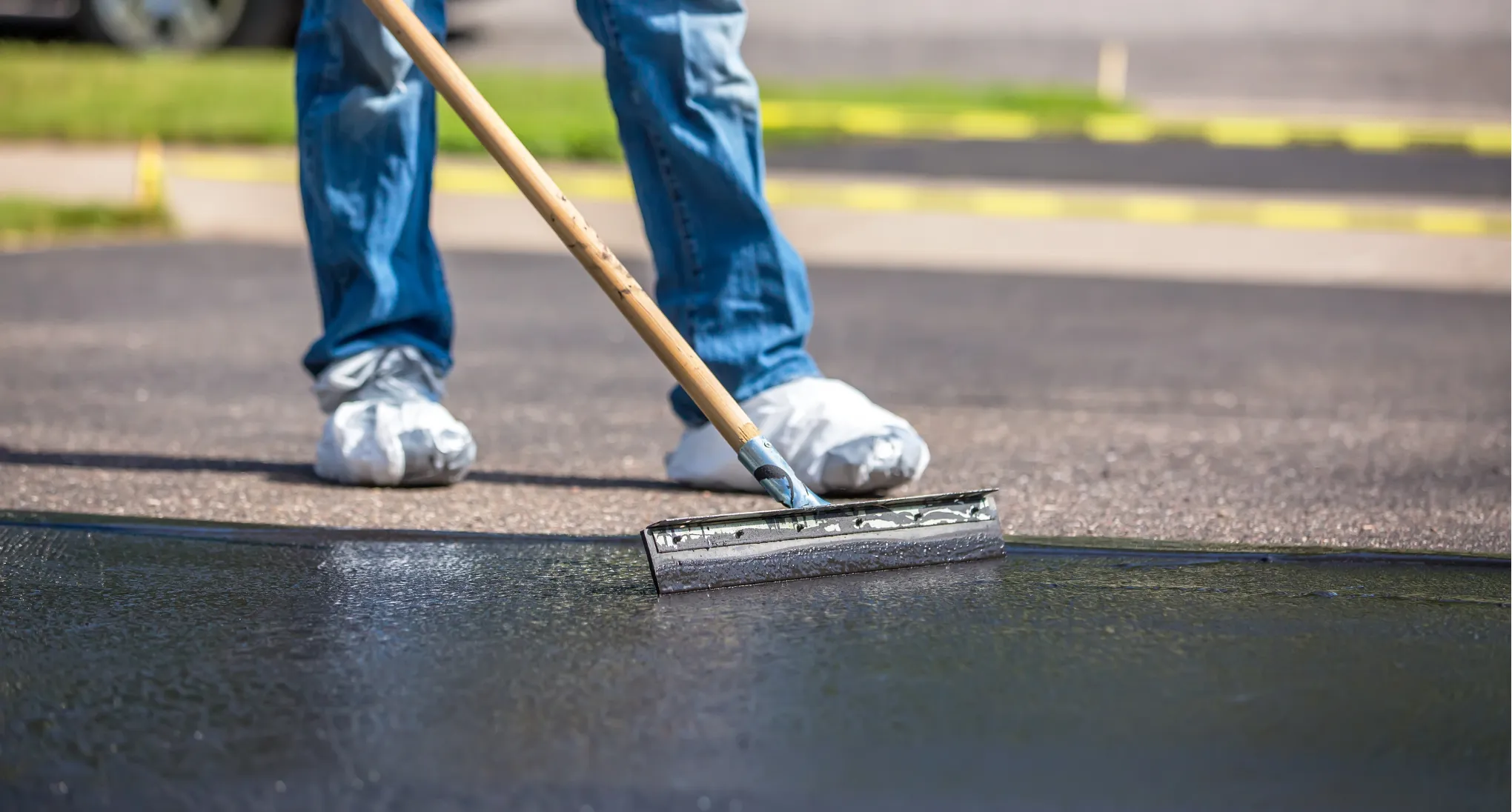 sealcoating-1 Person spreading asphalt sealer on a driveway with a squeegee, wearing jeans and protective shoe covers.