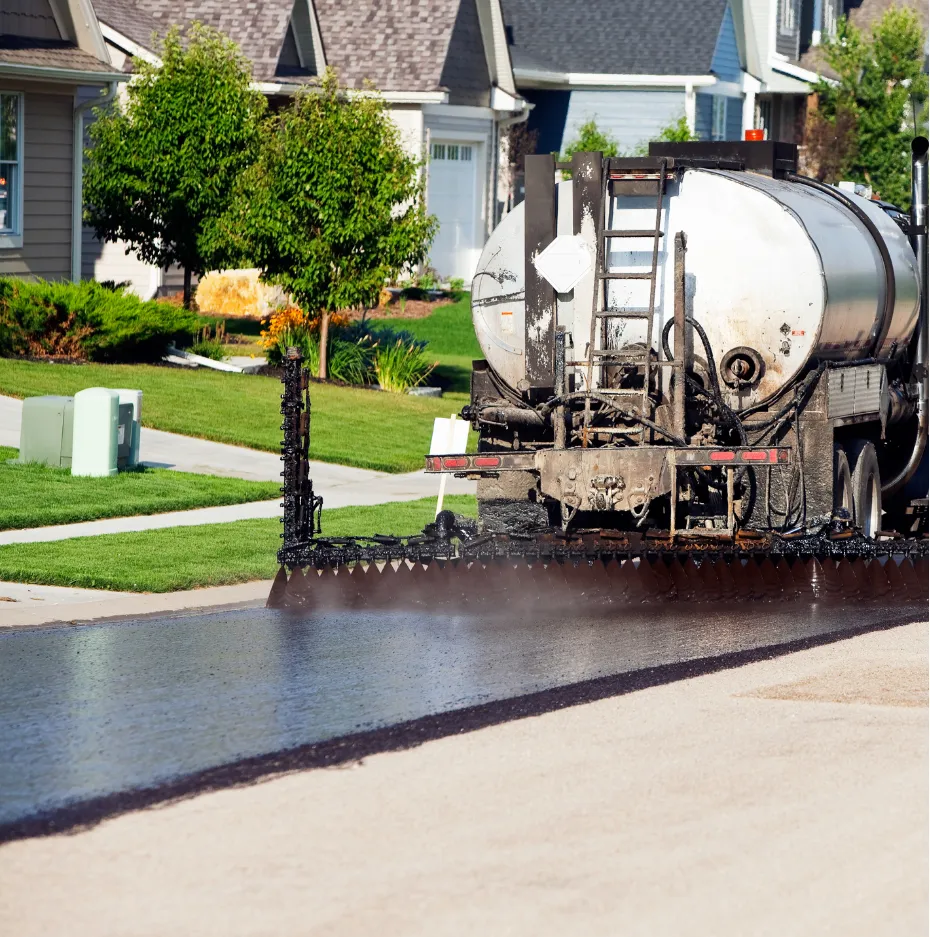Truck spraying asphalt sealant on a residential street, with houses and green lawns in the background.