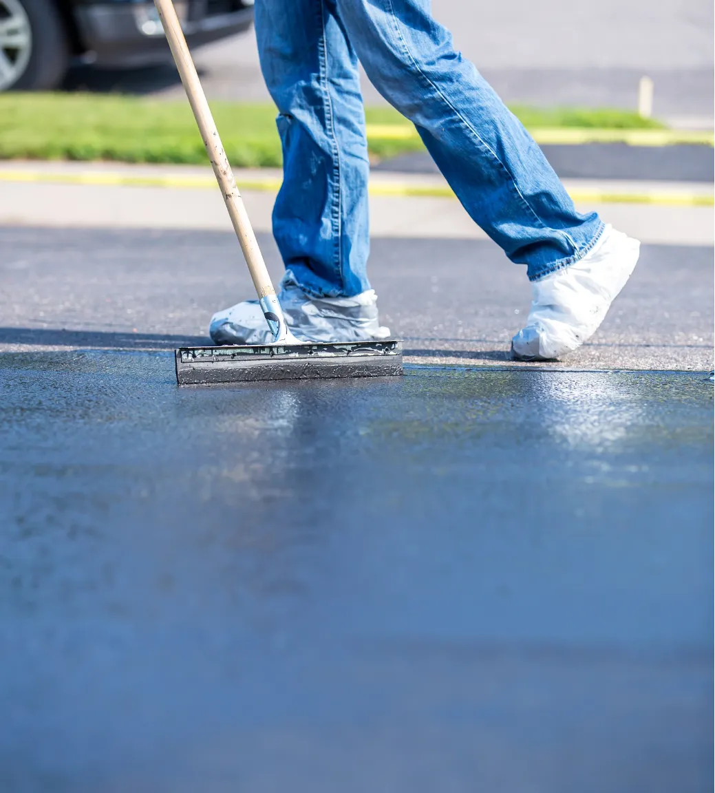 Person applying a fresh coat of sealant on a driveway with a large squeegee, wearing blue jeans and protective shoe covers.