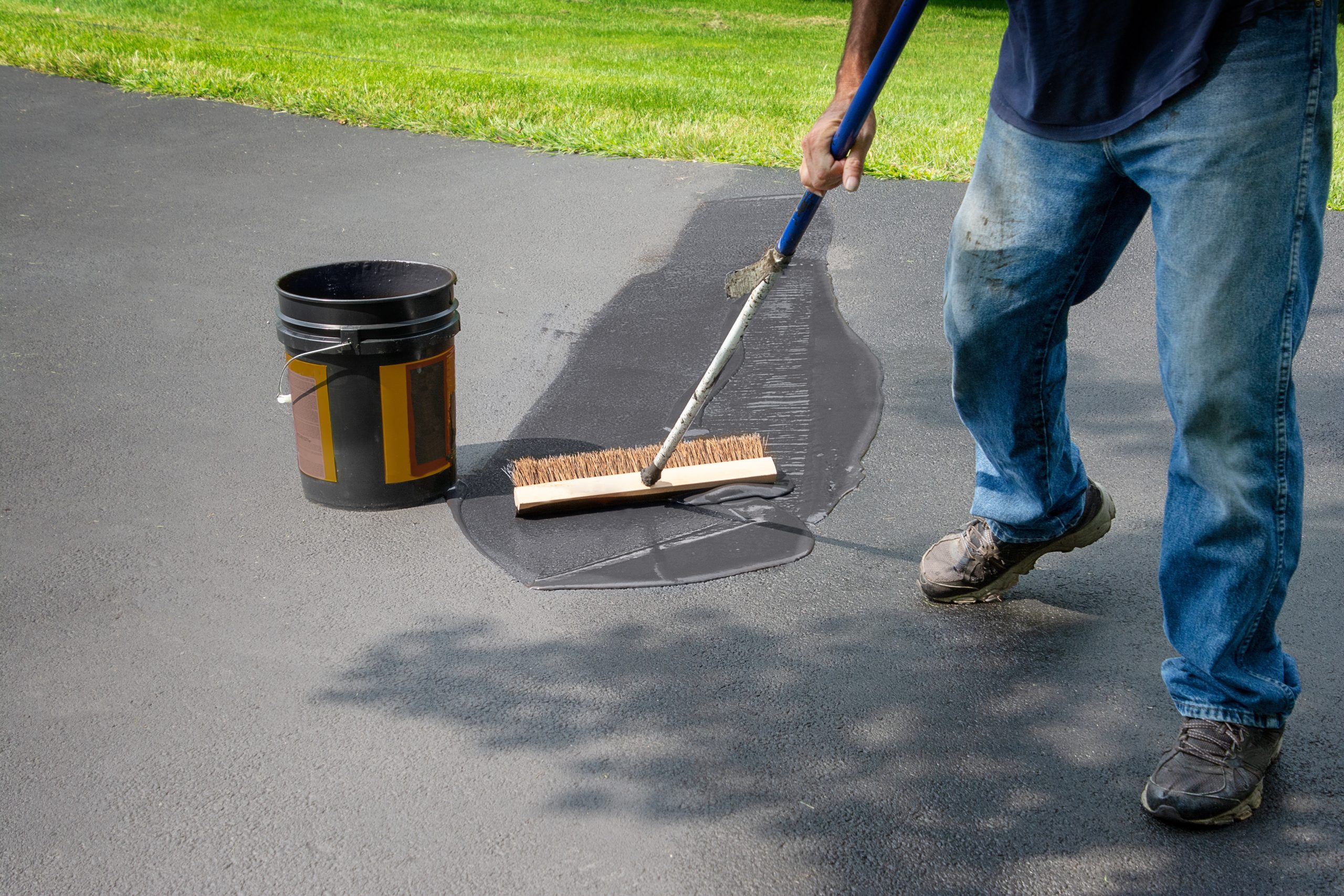 Homeowner spreads blacktop asphalt sealant on driveway Person sealing a driveway with a brush and bucket on a sunny day, grass in the background.