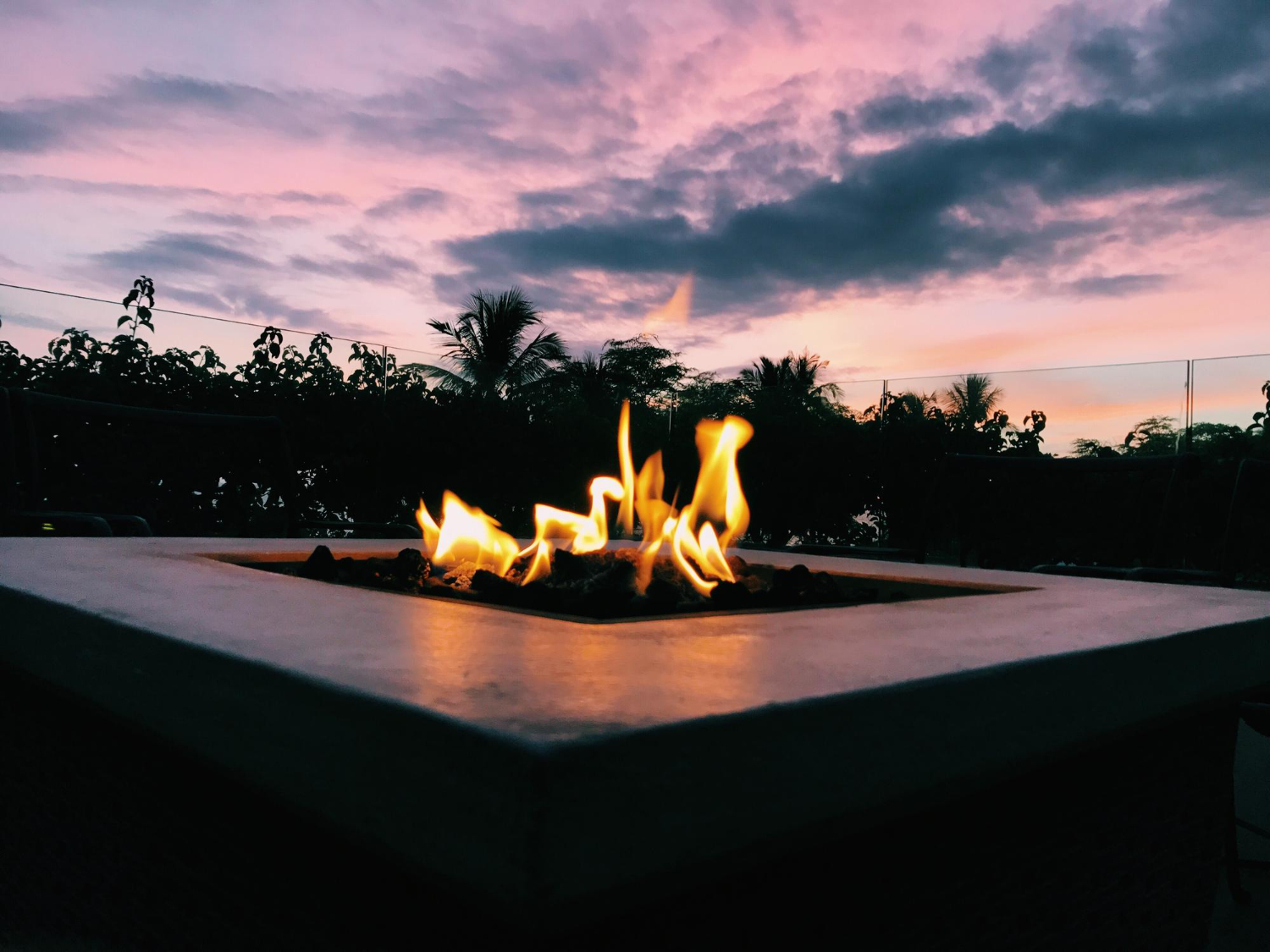 A fire pit with flames burning at sunset, trees and a colorful sky in the background.