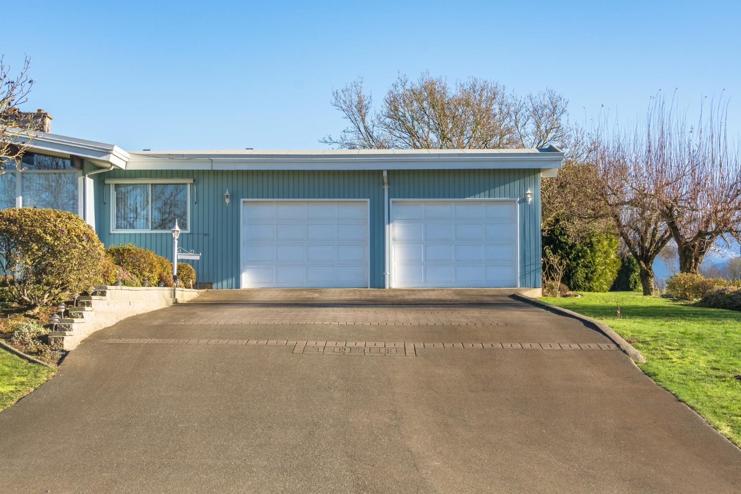 1975 Blue house with two white garage doors, wide driveway, and trees in the background under clear blue sky.