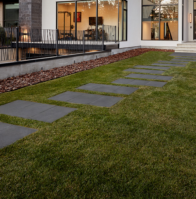 Expo_Dimensions_2023_03 Stepping stones lead across a green lawn to a modern house with large windows and outdoor seating.