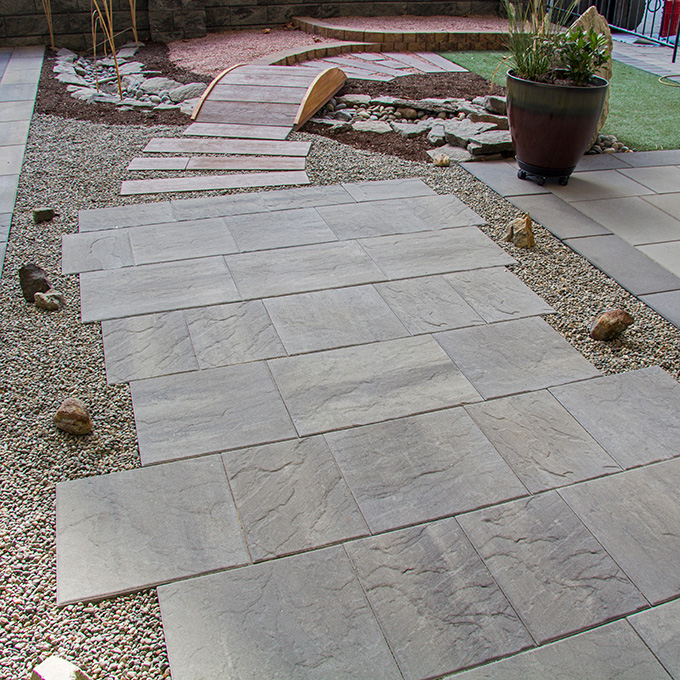 Expo_NewYorkStone_2023_01 Stone tile walkway over gravel, leading to a small wooden bridge with a planter nearby in a landscaped garden.