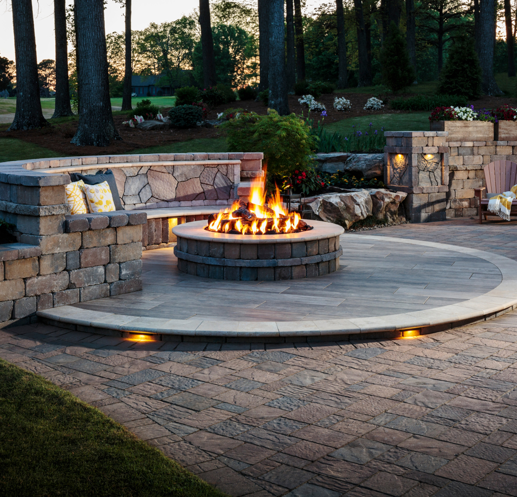 Inspiration_Fire-Pits_Image2 Stone patio with a round fire pit, built-in seating, and surrounding trees at dusk.