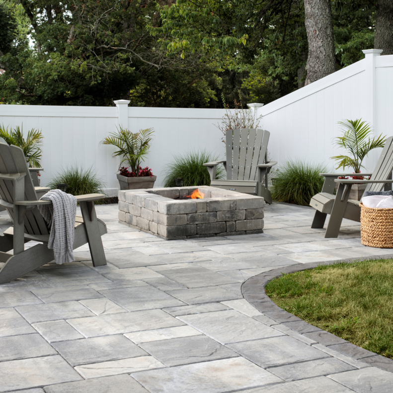 Inspiration_Fire-Pits_Image5 Gray Adirondack chairs surround a stone fire pit on a paved patio with plants and a white fence.
