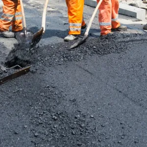 Workers in orange pants are spreading fresh asphalt on a road with shovels.