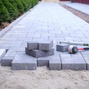 Gray paving stones being laid on sand, with tools and stacked bricks on a partially finished walkway.