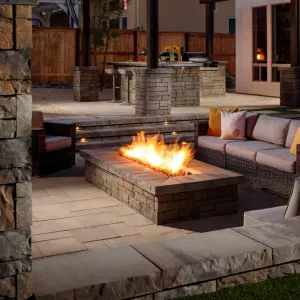Rectangular stone fire pit with flames on a patio, surrounded by cushioned outdoor sofas at night.