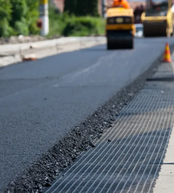 overlays-1 Fresh asphalt being paved on a road with construction equipment and traffic cones in the background.