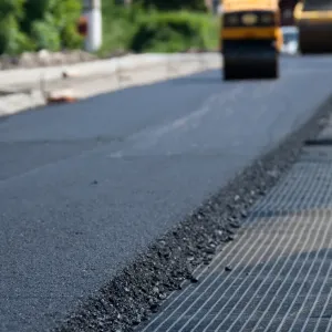 Close-up of fresh asphalt being laid on a road, with a road roller machine in the background.