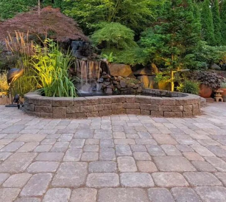 patios-2 Stone patio with raised pond, waterfall, lush greenery, and landscape lighting in a peaceful garden setting.