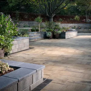 Spacious patio with stone pavers, raised flower beds, and potted plants under a large tree.