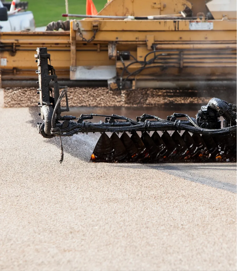 tar-chip-1 Close-up of machinery applying fresh asphalt to a road surface during construction.