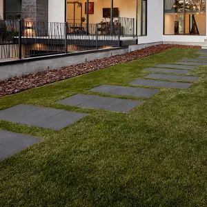 Modern backyard with square stepping stones on green grass, leading to a patio and a building with large windows.