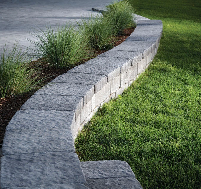walls-1-1 Gray stone retaining wall curves between a grassy lawn and a paved area with ornamental grasses planted behind.