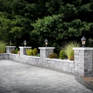 Stone driveway wall with columns, lantern lights, and greenery in front of dense pine trees.