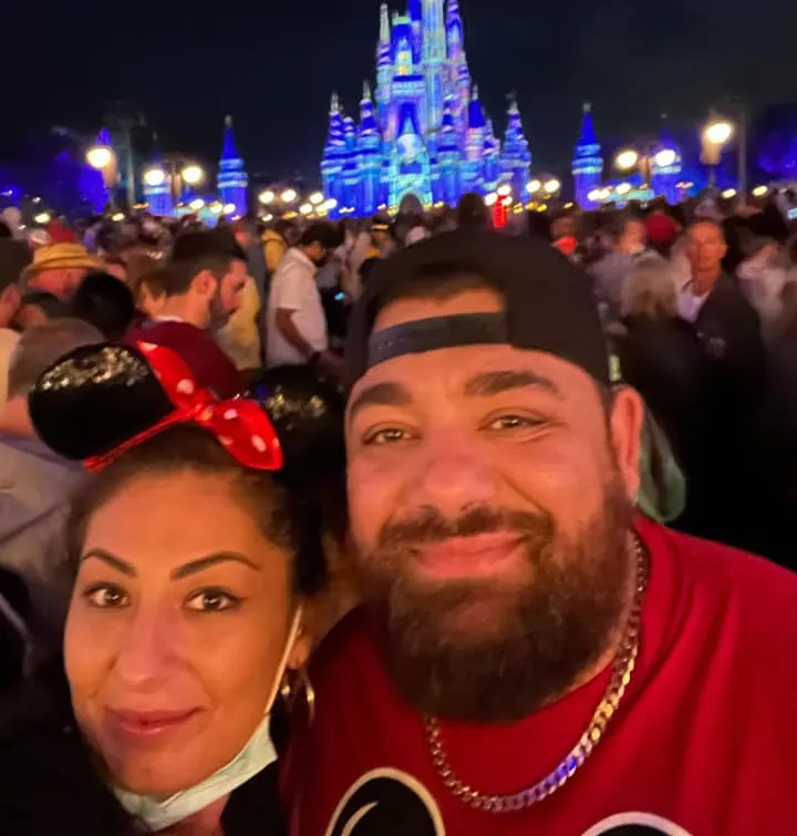A smiling couple at night in front of a brightly lit castle, surrounded by a crowd at a theme park.
