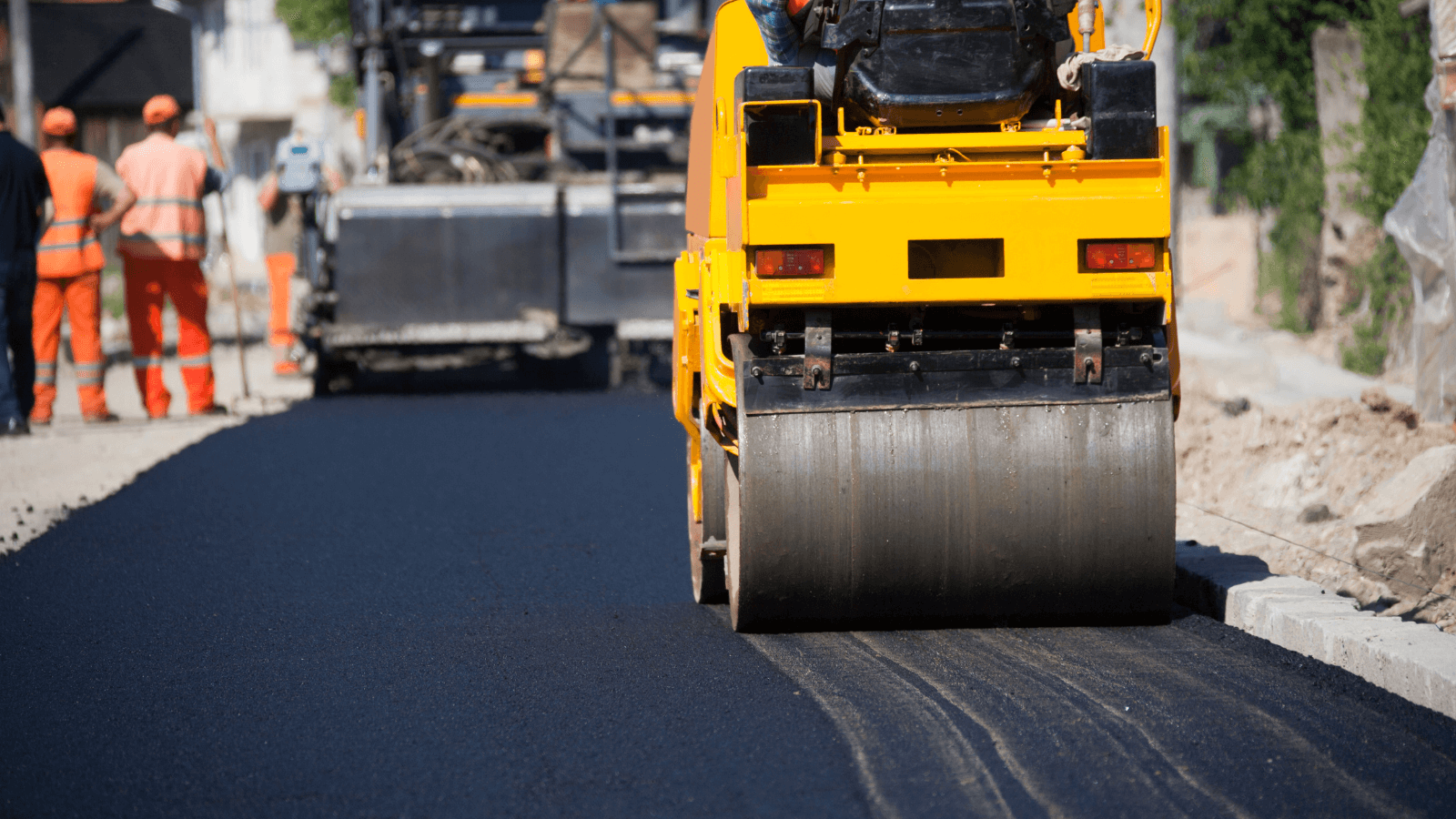 A yellow road roller compacts fresh asphalt as workers in orange uniforms work in the background.